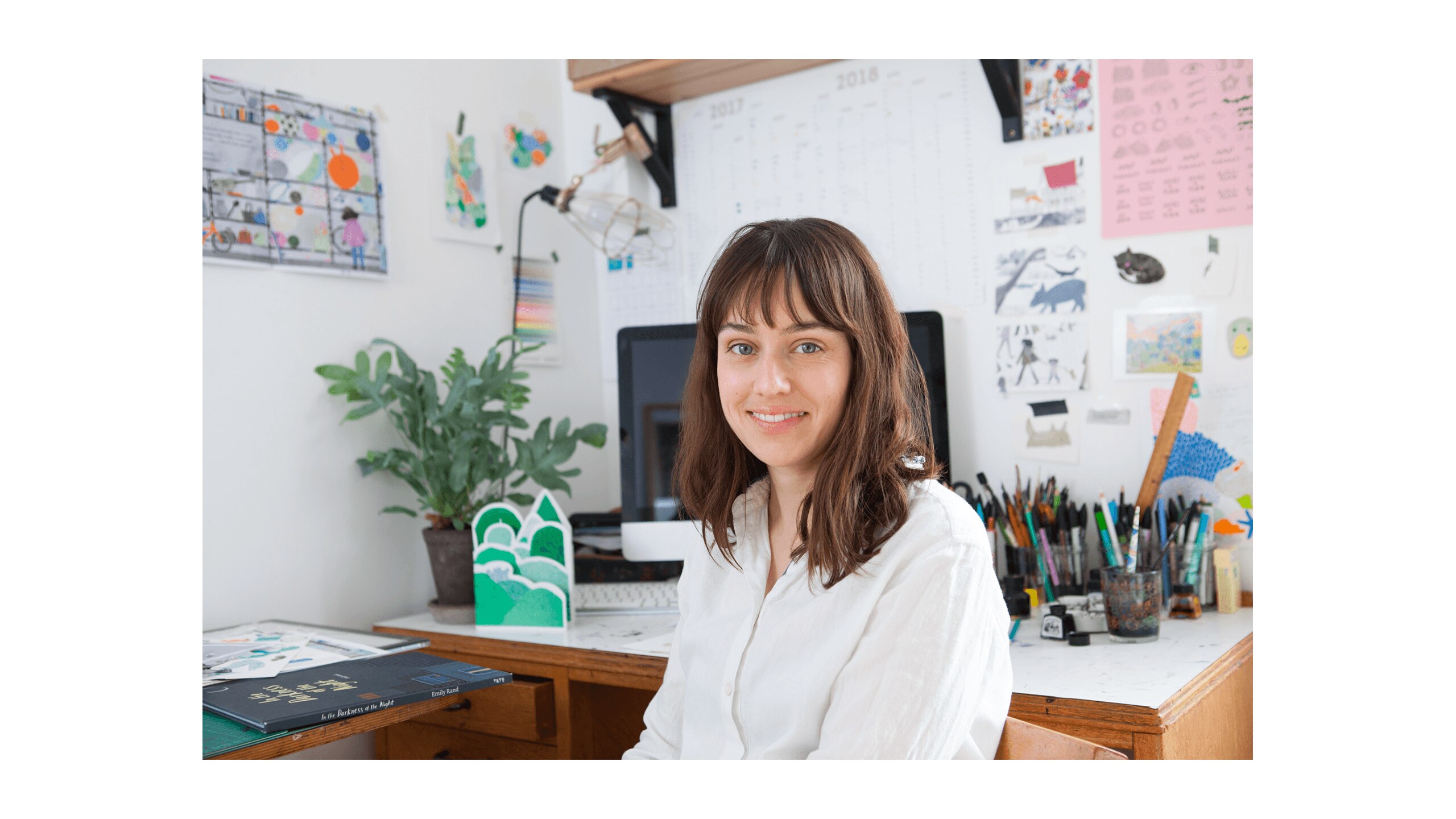 Photo of artist Emily Rand sat at her desk with a plant and a lot of pens.