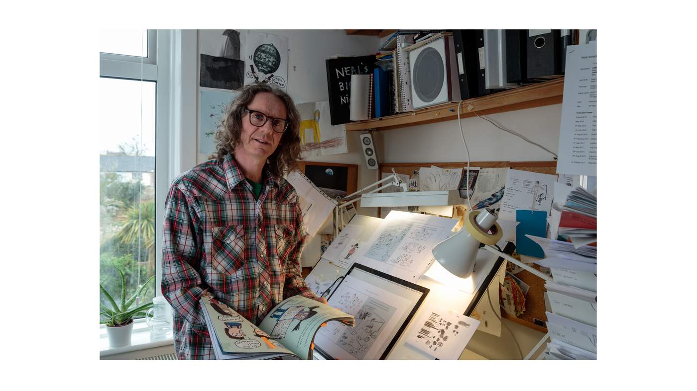 Artist Neal Layton poses holding a book of illustrations near his desk.